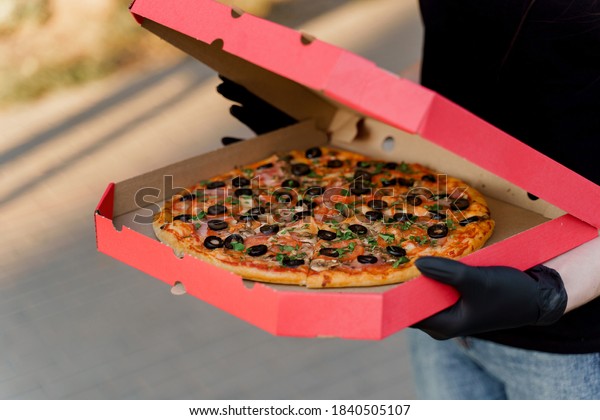 Food courier in black medical gloves holds red\
box with pizza from pizzeria. Food delivery at quarantine\
coronavirus covd-19\
period.