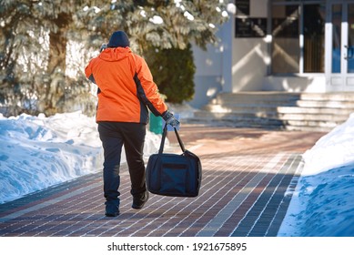 Food Courier In Orange Uniform Delivers Food Orders To Offices And Homes In Winter. Man Delivering Online Food Orders. Pizza Delivery Man Carries Bag In Hand. Delivery Service Around The Clock