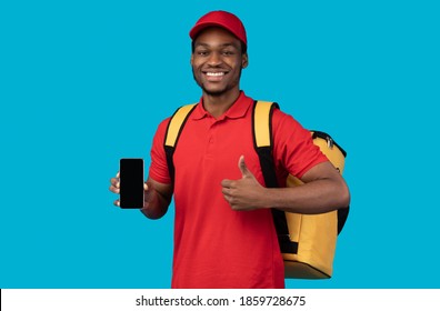 Food Delivery. African American Courier Man Holding Mobile Phone With Blank Screen For Mockup, Wearing Red Cap, Uniform And Thermo Backpack Bag, Showing Thumbs Up Isolated On Blue Background