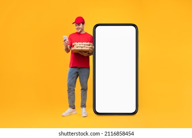 Food Delivery Application. Courier Guy Near Big Smartphone Holding Pizza Boxes And Using Mobile Phone Standing On Yellow Studio Background, Wearing Red Uniform. Mockup, Full Length