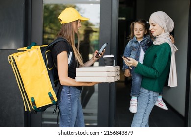 Food delivery to a beautiful muslim woman with little daughter. A young courier delivers an order to a customer. Order food and fast food online. Islamic woman pays by credit card, online payment.