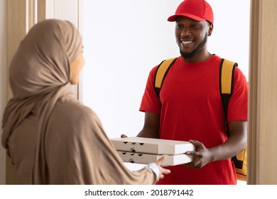 Food Delivery. Cheerful Black Male Courier Standing And Holding Pizza Boxes Smiling To Muslim Customer Lady In Doors Of Her Home, Giving Order. Selective Focus. Cafe And Restaurant Shipping Service
