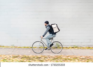 Food Delivery Courier Riding Fast In A Motion Blur Background Due To His High Speed. He Is Riding A Vintage Bike By A White Background Wall That Is Perfect For Copy Writing. Concept: Food Delivery