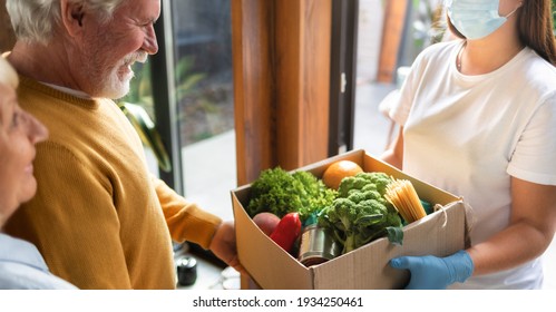 Food delivery or donation concept. Young woman in gloves and protective face mask delivers a box of food elderly couple in home - Powered by Shutterstock