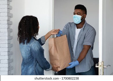 Food delivery during coronavirus. Black courier guy wearing medical mask delivering grocery order to young woman's home