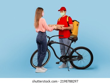 Food Delivery. Male Courier Delivering Pizza Boxes To Woman Standing Near Bike Wearing Face Mask Posing On Blue Background, Studio Shot. Transportation Service Concept. Full Length