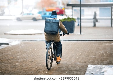 Food delivery man riding bicycle on snowy city street with large black backpack. Delivery boy on bike with thermal bag deliver pizza from restaurants during snow storm - Powered by Shutterstock