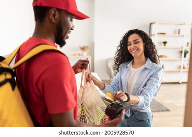 Food Delivery Service And Cashless Payment. Smiling Lady Paying With Debit Card For Groceries Receiving String Net Bag With Products Supplies From Supermarket, Deliveryman Standing At Door