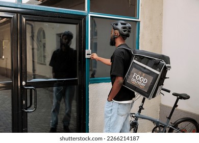 Food delivery service courier ringing office building doorbell, waiting for customer outdoors. African american man delivering restaurant takeaway meal, standing in front of door - Powered by Shutterstock