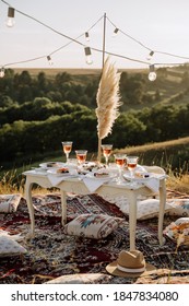 Food and Drink Glasses on Picnic Wooden Table. Beverage, Dessert on Plates and Dried Branch Decorate on Desk. Comfortable Pillows and Hat Carpet, Lighting Lamps at Nature. Forest on Background