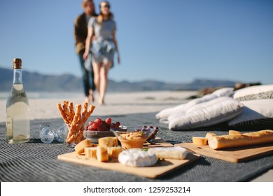 Food and drink on the blanket at the beach with couple walking towards it. Picnic setting on the beach with man and woman in background. - Powered by Shutterstock