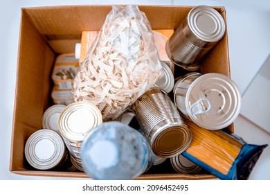 Food For The Endangered And Hungry During Crises. Donating And Fight Hunger By Giving Preserved And Caned Food. Top View On Groceries In Cardboard Box