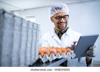 Food Factory Technologist In White Coat Hairnet And Hygienic Gloves Controlling Eggs Production At The Food Processing Plant On Tablet Computer.