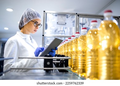 Food factory worker in sterile white uniform and hairnet using digital tablet while bottled refined oil is being produced on automated conveyor machine. - Powered by Shutterstock