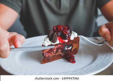 Food, Junk-food, Culinary, Baking And Holidays Concept - Close Up Of Male Hands Eating Chocolate Cherry Cake With Fork And Sitting At Wooden Table.tasty Dessert.chocolate Cake Slice On White Plate.