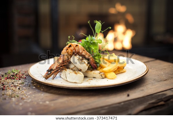 Food - Main course gourmet\
seafood - Delicious grilled king prawn with chicken fillet and\
homemade french fries served on a wooden table, fireplace on\
background