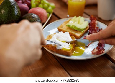 Food And People Concept - Hands Of Man Eating Toast With Pouched Egg, Avocado And Bacon With Fork And Knife