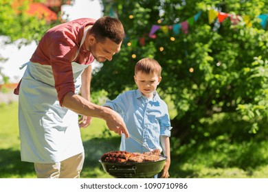 Food, People And Family Time Concept - Father And Son Cooking Meat On Barbecue Grill At Bbq Party In Summer Garden