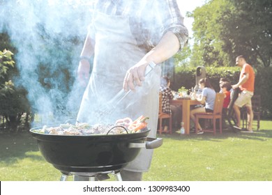 Food, People And Family Time Concept - Man Cooking Meat On Barbecue Grill At Summer Garden Party