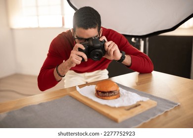 Food photographer handsome young middle eastern man wearing casual outfit and eyeglasses taking photos of delicious juicy meaty hamburger at photo studio, using dslr camera