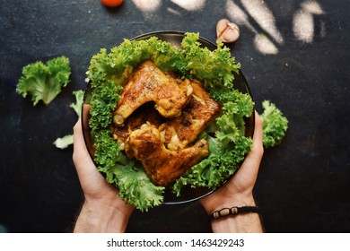 Food Photography. Chicken Wings With Lettuce In A Plate On A Black Background. Appetizing Grilled Meat, Barbecue. Tasty Dinner. A Man Holding A Plate Of Chicken Meat. Food In The Hand