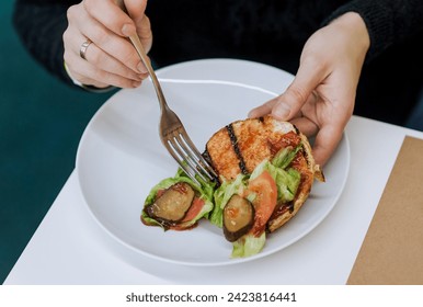 Food photography, fast food close-up. A woman with a fork in her hands eats a burger lying on a white plate with cucumbers, tomatoes, cabbage, meat, dividing it in half inside. - Powered by Shutterstock