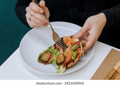 Food photography, fast food. A woman with a fork in her hands eats a burger with cucumbers, tomatoes, cabbage, meat lying on a white plate, dividing it in half inside. - Powered by Shutterstock