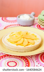 Food Photography Of A Mango Custard Tart (flan) On A Colourful Table Setting Of Yellow, Orange, Red, Pink And Green With A Ramekin Of Whipped Cream In The Background