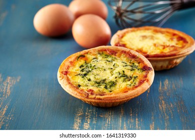 Food photography of mini quiches florentine (spinach) and lorraine (ham) tarts with eggs and a whisk on a rustic blue painted wooden background