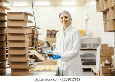 A food plant worker is collecting cookies while smiling at the camera.