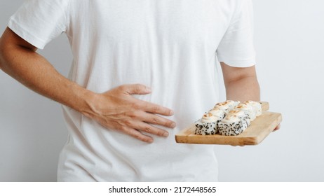 Food poisoning, indigestion, stomach pain, allergy to asian food concept. Close-up of sick man holding his belly and sushi on wooden board indoors.