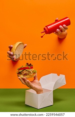 Food pop art photography. Female hand sticking out orange paper with ketchup over burger on hand sticking out food box. Concept of taste, creativity, art. Complementary colors. Copy space for ad, text