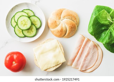 Food Products And Ingredients For Making Sandwich. Ham, Cheese, Burger Bun, Lettuce, Cucumber And Tomato As Recipe Flatlay On Marble Kitchen Table Background