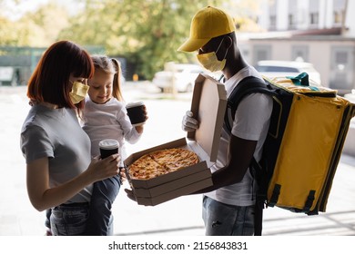 Food Shipping And People Concept - Happy Delivery African Man In Face Mask With Thermal Insulated Bag Giving Pizza Box And Coffee To Young Family Mother And Daughter Customers To Their Home.