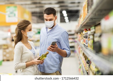 Food Shopping, Consumerism And Pandemic Concept - Happy Couple Wearing Face Protective Medical Masks For Protection From Virus Disease With Smartphone Buying Olive Oil At Grocery Store Or Supermarket