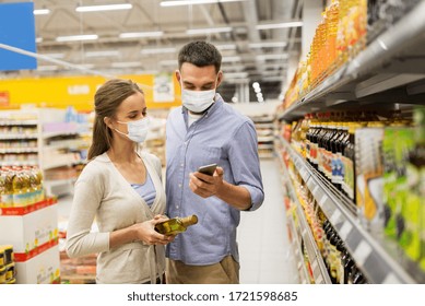 Food Shopping, Consumerism And Pandemic Concept - Happy Couple Wearing Face Protective Medical Masks For Protection From Virus Disease With Smartphone Buying Olive Oil At Grocery Store Or Supermarket