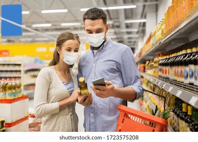 Food Shopping, Consumerism And Pandemic Concept - Happy Couple Wearing Face Protective Medical Masks For Protection From Virus Disease With Smartphone Buying Olive Oil At Grocery Store Or Supermarket