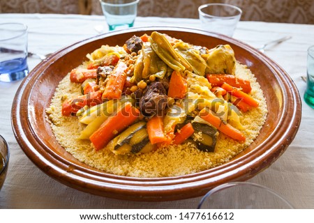 Food shot of a typical moroccan couscous served on a family size ceramics tagine plate surrounded by glasses, sauce, spoons and spices on a linens background. Semolina, vegetables and meat ingredients