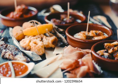 The food of Spain. An overhead photo of many different Spanish tapas, shot from above on a dark rustic texture. Jamon, cheese, wine, olives, croquettes, calamari rings, anchovies and more