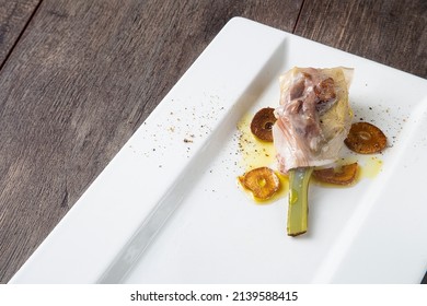 Food still life with artichoke and baked pork jowl with garlic slices on a white plate with wooden board background, with copy space.