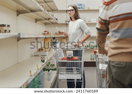 Food supply shortage and panic buying: worried woman doing grocery shopping at the supermarket and staring at the almost empty shelves