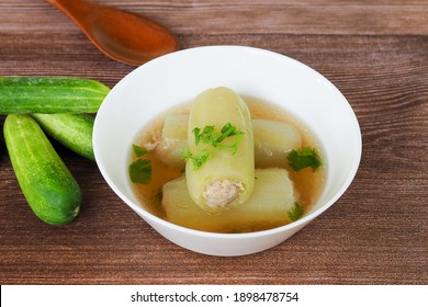 Food from Thailand Cucumber soup stuffed with minced pork in a white bowl is placed on a wooden table. And there is a wooden spoon, cucumber, garlic, spring onion, coriander placed next to the bowl.