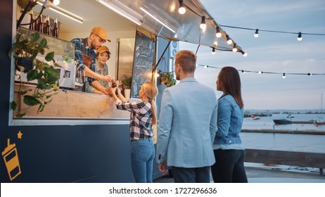 Food Truck Employee Hands Out Beef Burgers, Fries and Cold Drinks to Happy Hipster Customers. People are Eating Outside. Commercial Truck Selling Street Food in a Modern Place Near the Sea. - Powered by Shutterstock