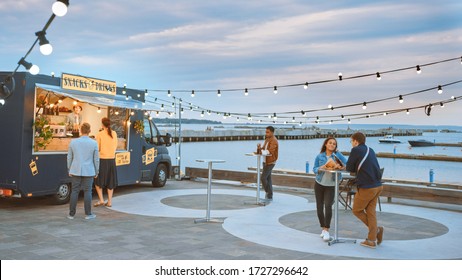 Food Truck Employee Hands Out Beef Burgers, Fries and Cold Drinks to Happy Hipster Customers. People are Eating at Tables Outside. Commercial Truck Selling Street Food in a Modern Place Near the Sea. - Powered by Shutterstock