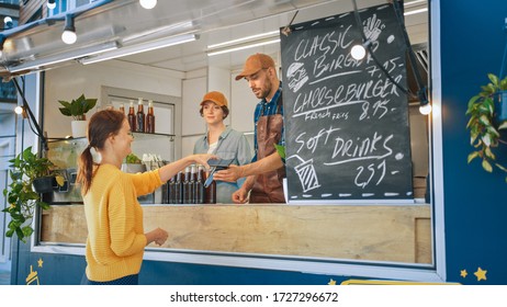 Food Truck Employee Hands Out a Freshly Made Burger to a Happy Young Female. Young Lady is Using NFC Mobile Payment to Pay for Food. Street Food Truck Selling Burgers in a Modern Hip Neighbourhood. - Powered by Shutterstock