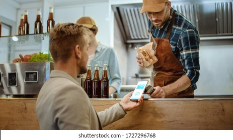 Food Truck Employee Hands Out a Freshly Made Burger to a Happy Young Male. Man in a Casual Suit is Using His Smartphone for NFC Mobile Payment Solution. Street Food Truck Selling Burgers. - Powered by Shutterstock