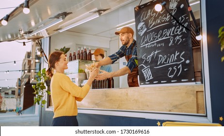 Food Truck Employee Hands Out A Freshly Made Gourmet Burger To A Happy Young Female. Young Man In A Suit Is Making An Order. Street Food Truck Selling Burgers In A Modern Hip Neighbourhood.