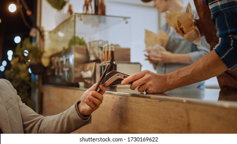 Food Truck Employee Hands Out a Freshly Made Burger to a Happy Young Male. Man in a Casual Suit is Using NFC Mobile Payment Solution. Street Food Truck Selling Burgers in a Modern Hip Neighbourhood. - Powered by Shutterstock