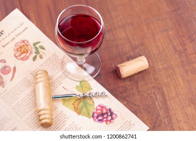 Food Writing Or Restaurant Critique Concept. Glass Of Red Wine With An Old Newspaper, A Vintage Corkscrew And A Cork In The Blurred Background, On A Wooden Table With A Place For Text. Selective Focus