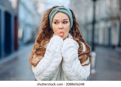 Fooling around on camera. A young funny girl in a white knitted suit and a blue headband poses droll on the street. Eyes averted and tongue protruding. Blurry background. The concept of silly photos. - Shutterstock ID 2088656248